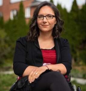 Photo of Dr. Marjorie Aunos. She is outside sitting in her wheelchair with a bright red top and black jacket.
